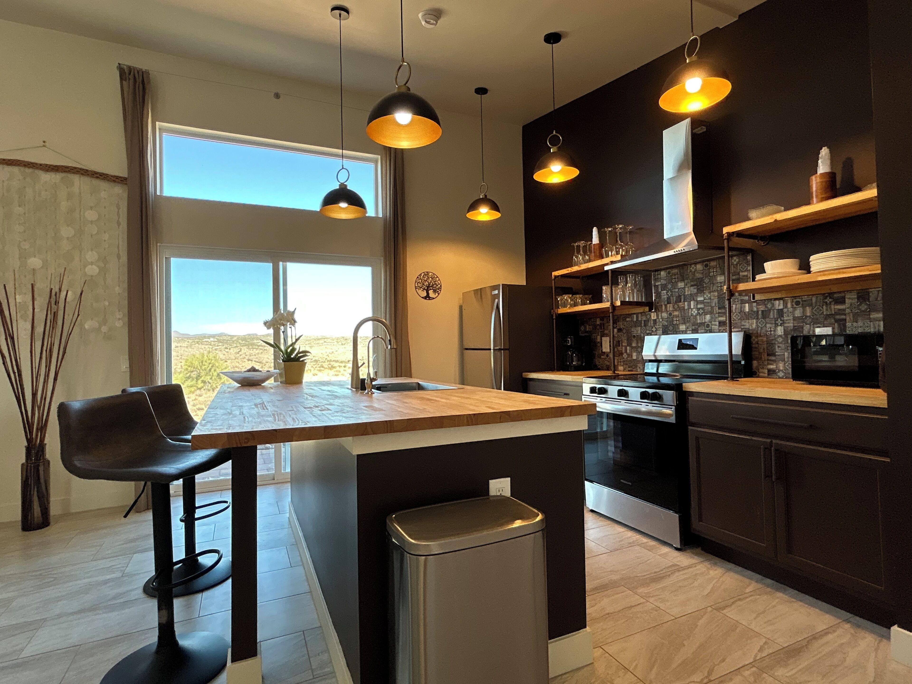 A modern kitchen with a central island, stainless steel appliances, pendant lighting, and a view of the desert through the window.