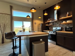 A modern kitchen with a central island, stainless steel appliances, pendant lighting, and a view of the desert through the window.
