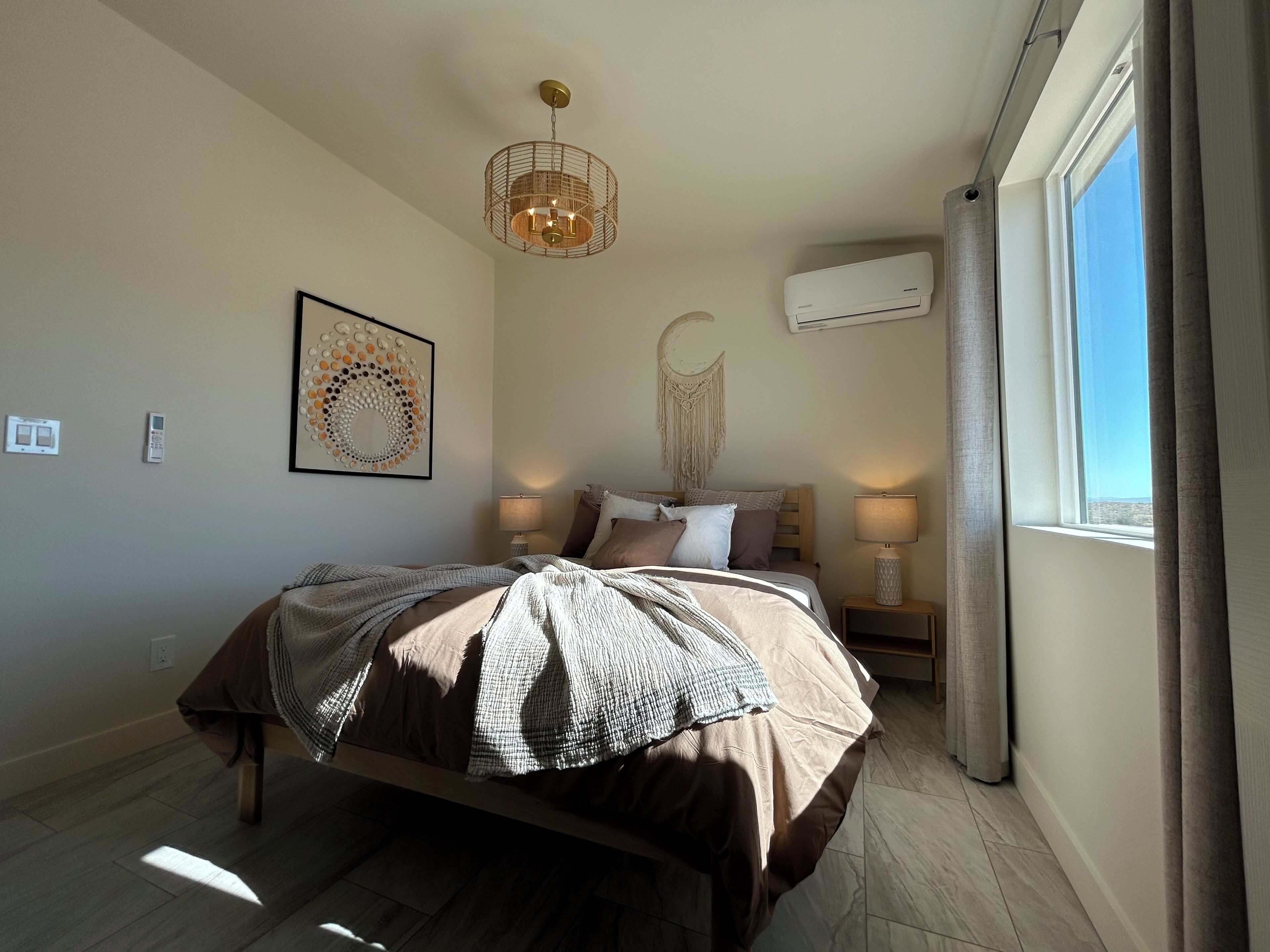 A bedroom with a bed, bedside lamps, a wall-mounted air conditioner, and a window with curtains.