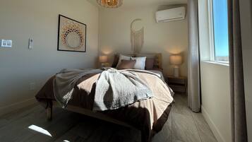 A bedroom with a bed, bedside lamps, a wall-mounted air conditioner, and a window with curtains.
