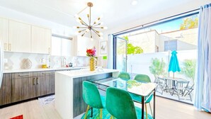Une cuisine moderne dotée d’un îlot central, d’un coin repas avec une table en verre et des chaises vertes, et offrant une vue sur l’extérieur grâce à de grandes fenêtres.