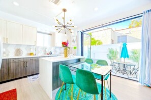 A modern kitchen with a central island, a dining area with a glass table and green chairs, and a view of the outdoors through large windows.
