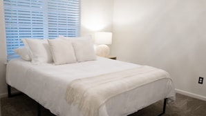 Une chambre à coucher avec un lit, du linge de lit blanc, une table de chevet avec une lampe et une fenêtre avec des stores.