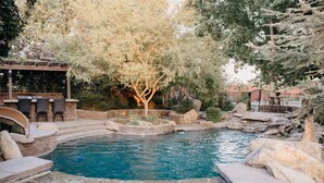 Une piscine avec un jacuzzi, entourée de rochers et d’arbres, avec un espace salon extérieur et une pergola.