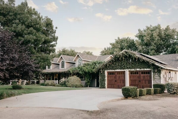 A two-story house with a covered garage, surrounded by a well-maintained lawn and mature trees. - Rustic Chateau/Estate -Pool, Spa, Tennis Court, and Quiet, Peaceful Surroundings (Del Rey)