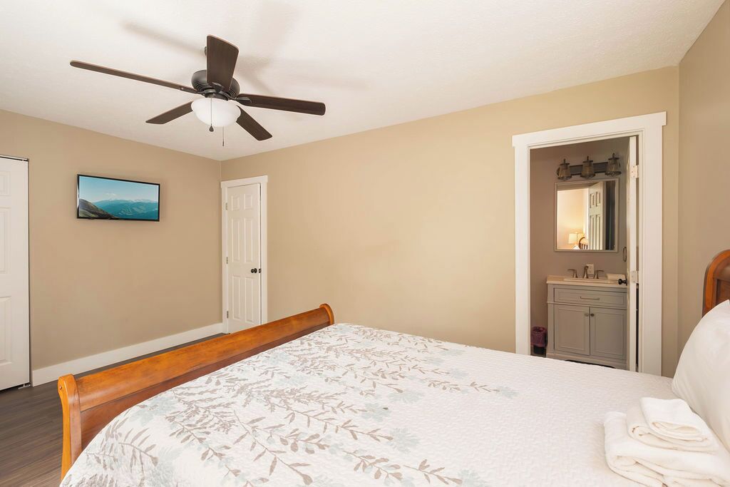 Ein Schlafzimmer mit Bett, Deckenventilator, Tür, Badezimmer mit Waschbecken und Spiegel sowie einem Fernseher an der Wand.