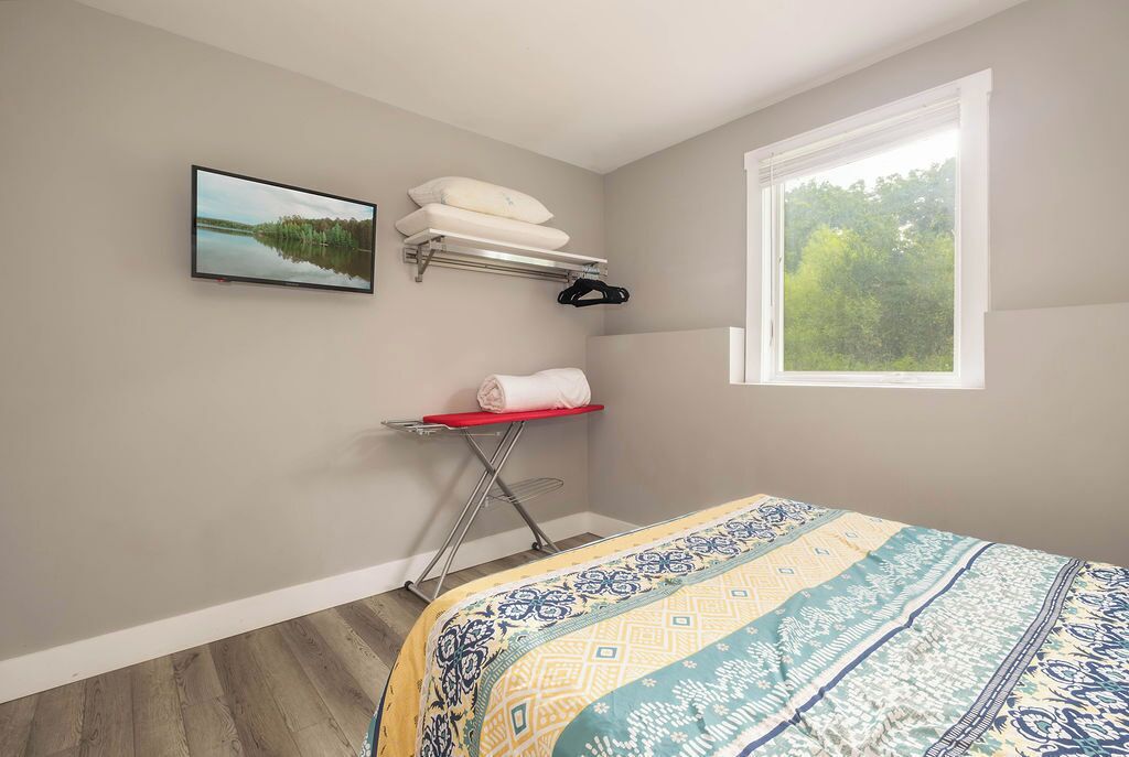 A bedroom with a bed, a window with a view of trees, a wall-mounted TV, and a shelf with towels and a hanger.