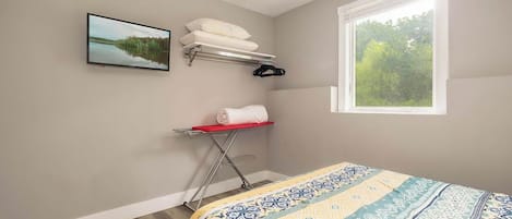 A bedroom with a bed, a window with a view of trees, a wall-mounted TV, and a shelf with towels and a hanger.