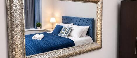 Un dormitorio con un espejo grande y ornamentado, una cama con ropa de cama azul, una mesita de noche con una lámpara y un armario oscuro.