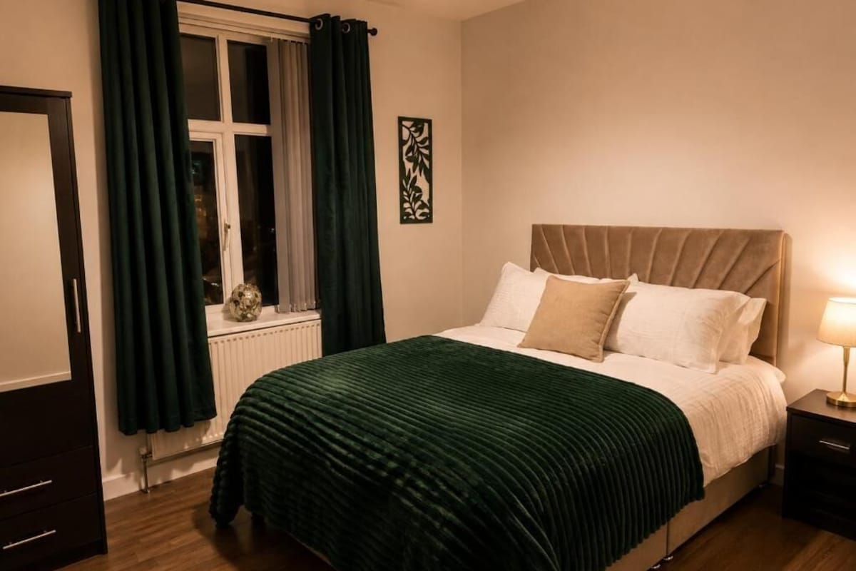 A bedroom with a bed, a nightstand, a window with curtains, and a radiator.