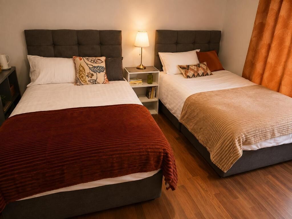 A hotel room with two beds, each with a different colored blanket, a bedside table with a lamp, and a nightstand with a cup.