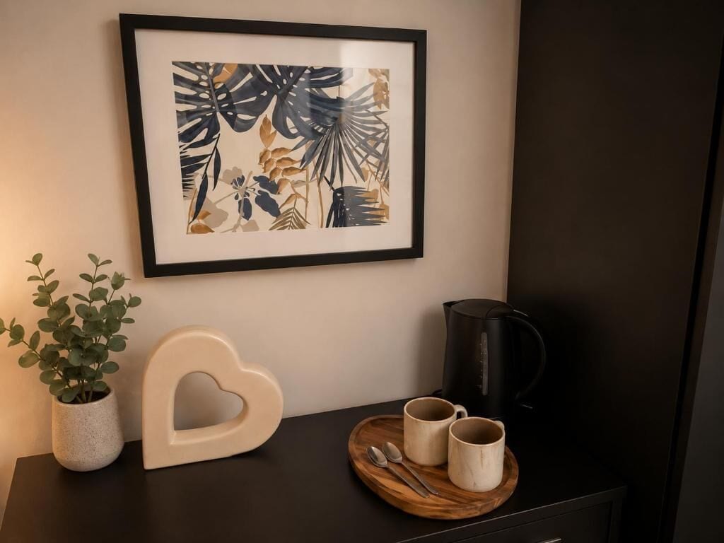 Ein gerahmtes Kunstwerk, ein herzförmiger Gegenstand, eine Kaffeemaschine und ein Tablett mit Tassen auf einer dunklen Fläche.