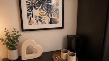 Una obra de arte en marco, un objeto con forma de corazón, una cafetera y una bandeja con tazas sobre una superficie oscura.