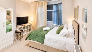 Un dormitorio con cama, televisor, ventana con cortinas y estante fijado a la pared.