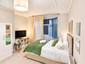 A bedroom with a bed, a television, a window with curtains, and a wall-mounted shelf. - Mauldeth Road Guest House (Manchester)