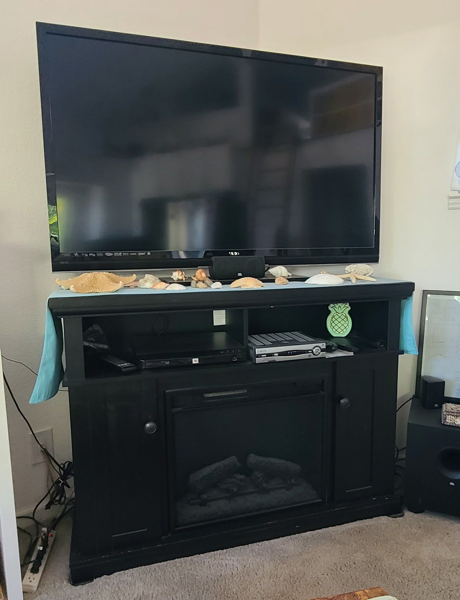 Un meuble de rangement noir pour appareils électroniques, avec un téléviseur à écran plat fixé au-dessus, une étagère ornée d’objets décoratifs et un petit meuble en dessous.