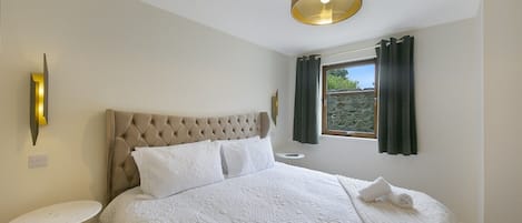 Un dormitorio con cabecera capitonada, una cama grande con ropa de cama blanca, una mesita de noche, una lámpara y una ventana con cortinas.