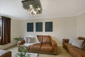 A living room with a brown leather sofa, two side chairs, a coffee table, and a modern chandelier. There are three abstract paintings on the wall. - Leslie Lodge - Grampian Lettings (Inverurie)