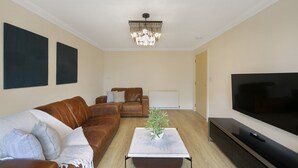 A living room with a brown leather sofa, a white coffee table with a plant, a large flat-screen TV, and a chandelier. - Leslie Lodge - Grampian Lettings (Inverurie)
