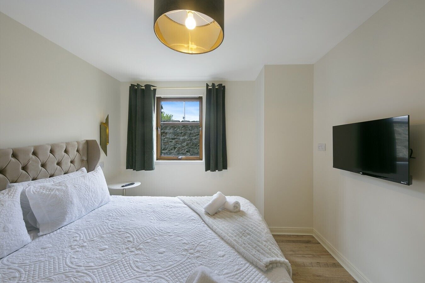 A hotel room with a bed, a small bedside table, a window with curtains, a wall-mounted TV, and a ceiling light fixture.