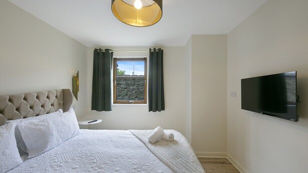 A hotel room with a bed, a small bedside table, a window with curtains, a wall-mounted TV, and a ceiling light fixture. - Leslie Lodge - Grampian Lettings (Inverurie)