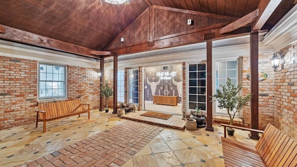 A spacious indoor area with a wooden ceiling, brick walls, and a stone floor. There are benches and potted plants.