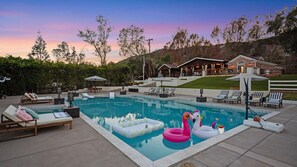 A swimming pool with lounge chairs and inflatable toys, surrounded by a landscaped area and a building in the background.