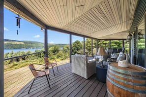 A covered outdoor deck with wicker furniture, a wooden barrel, and a view of a lake and hills. - Eagle's Landing | Cozy, Pet-Friendly Getaway (Hammondsport)