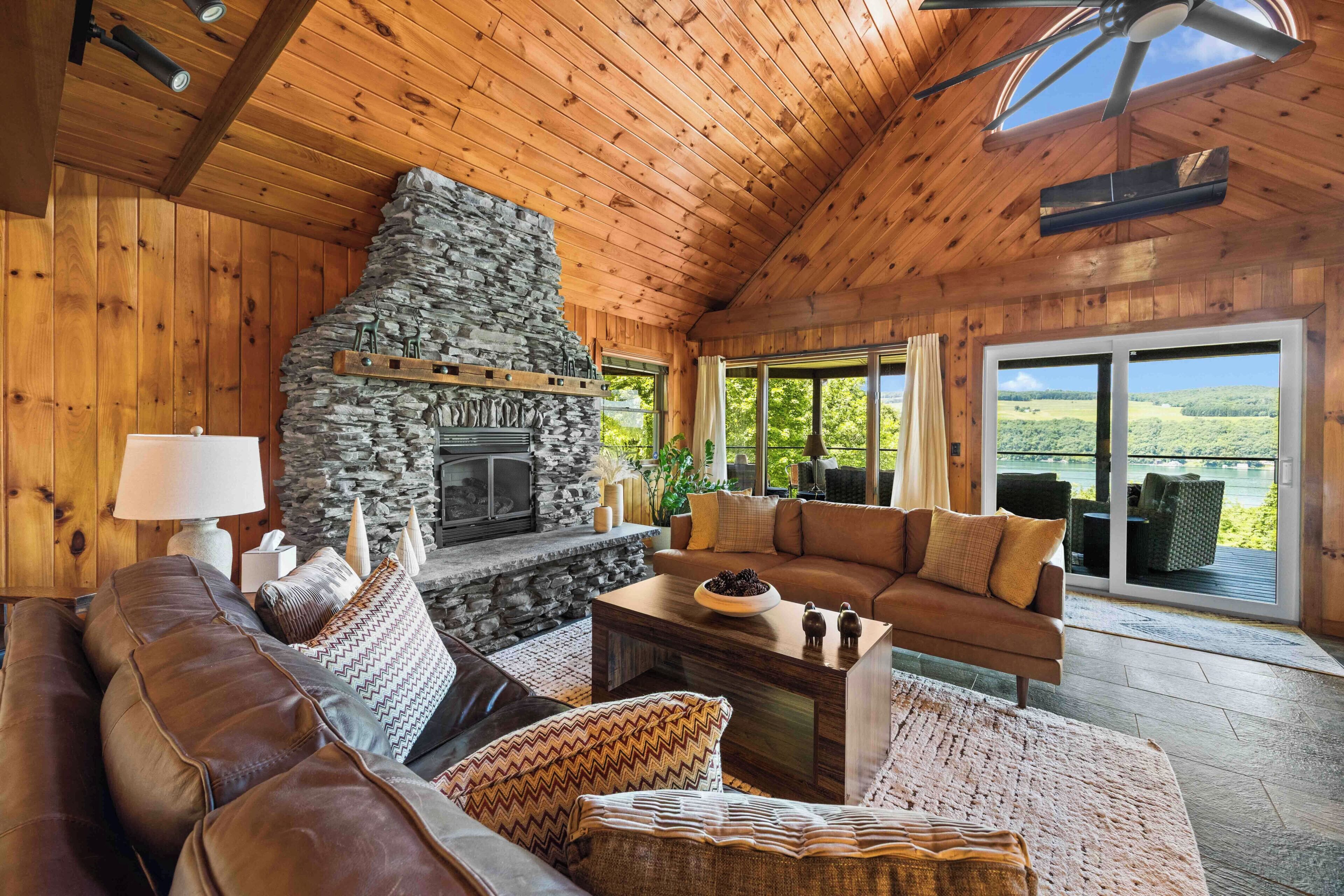 A cozy living room with a stone fireplace, wooden ceiling, and large windows offering a view of the outdoors.