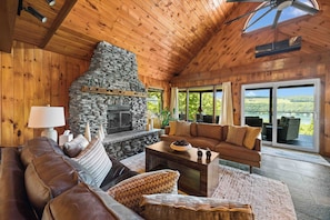 A cozy living room with a stone fireplace, wooden ceiling, and large windows offering a view of the outdoors. - Eagle's Landing | Cozy, Pet-Friendly Getaway (Hammondsport)