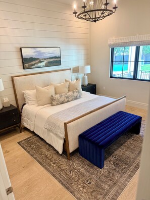 A bedroom with a large bed, a nightstand, a bedside table, a lamp, a window with a view, and a chandelier.