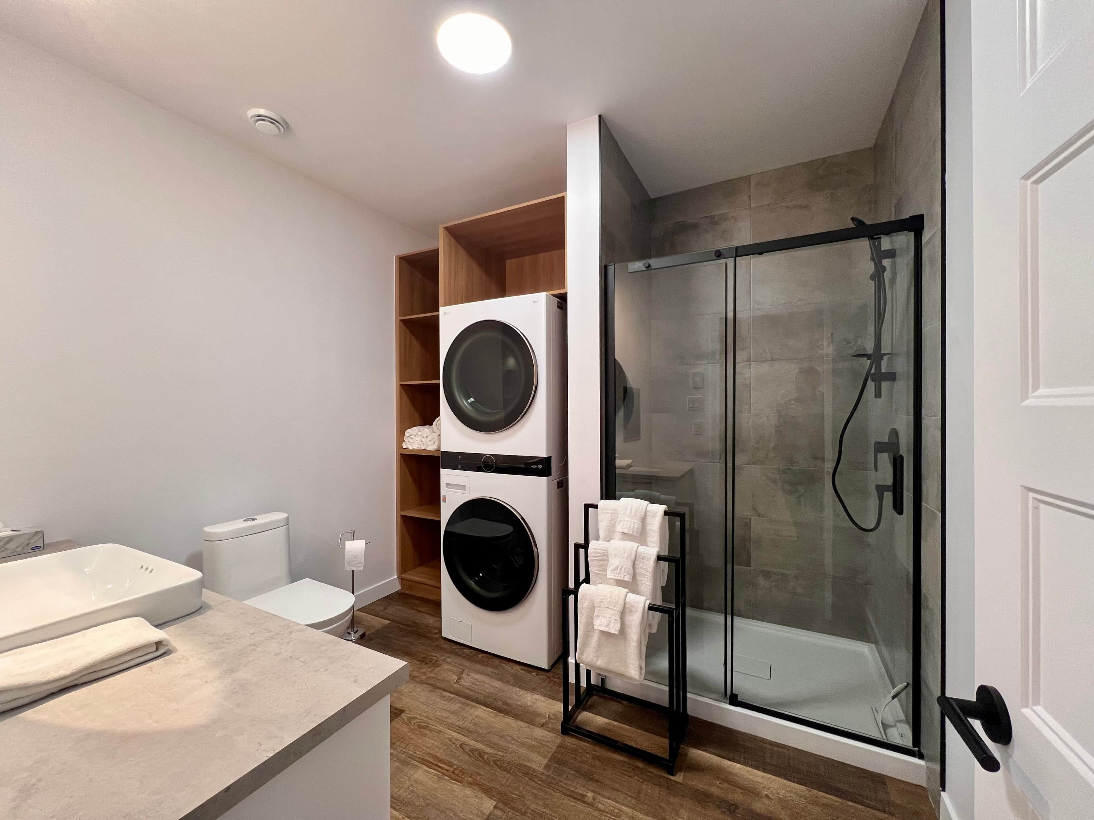 A modern bathroom with a glass-enclosed shower, a built-in washing machine, and a wooden shelving unit.