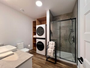 A modern bathroom with a glass-enclosed shower, a built-in washing machine, and a wooden shelving unit.