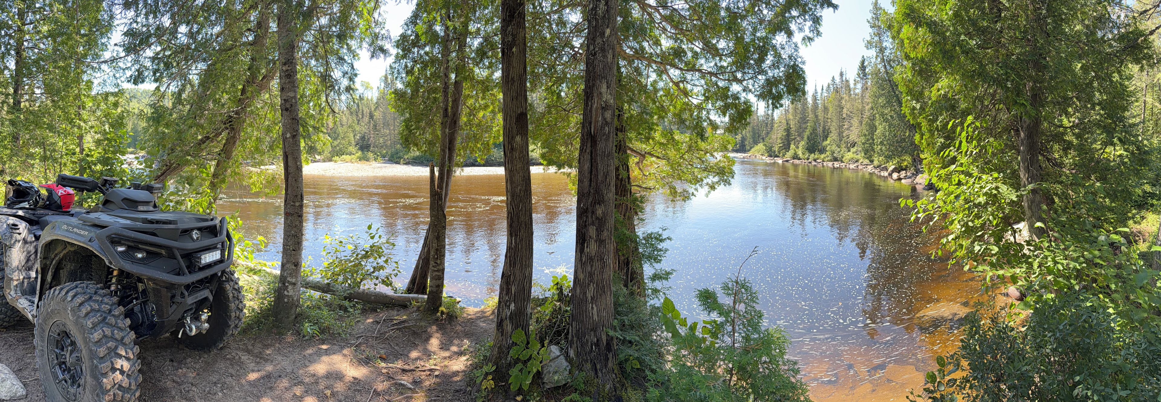 Une rivière bordée d’une forêt dense, avec un véhicule tout-terrain garé sur la rive gauche.