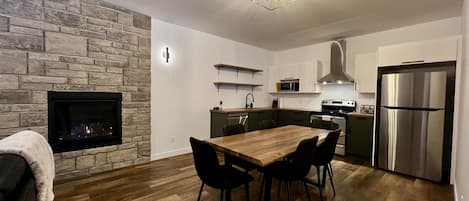 A modern kitchen with a stone wall, wooden flooring, a dining table with chairs, a built-in stove, and a stainless steel refrigerator.
