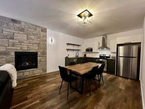 Una cocina moderna con pared de piedra, suelo de madera, una mesa de comedor con sillas, estufa integrada y refrigerador de acero inoxidable.