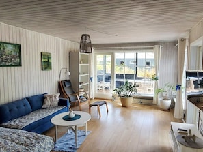 Sala de estar com sofá azul, mesa de centro branca, estante de livros e uma grande janela com vista para o exterior.