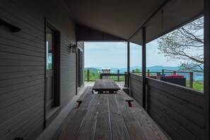 A covered porch with a wooden bench and a grill, overlooking a scenic view with mountains and a body of water. - Heated Pool, Hot Tub, Fire Pit & Theater - Asheville (Fairview)