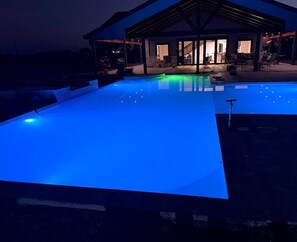 Une piscine dotée d’un système d’éclairage bleu, un patio couvert et un bâtiment avec une entrée ouverte.