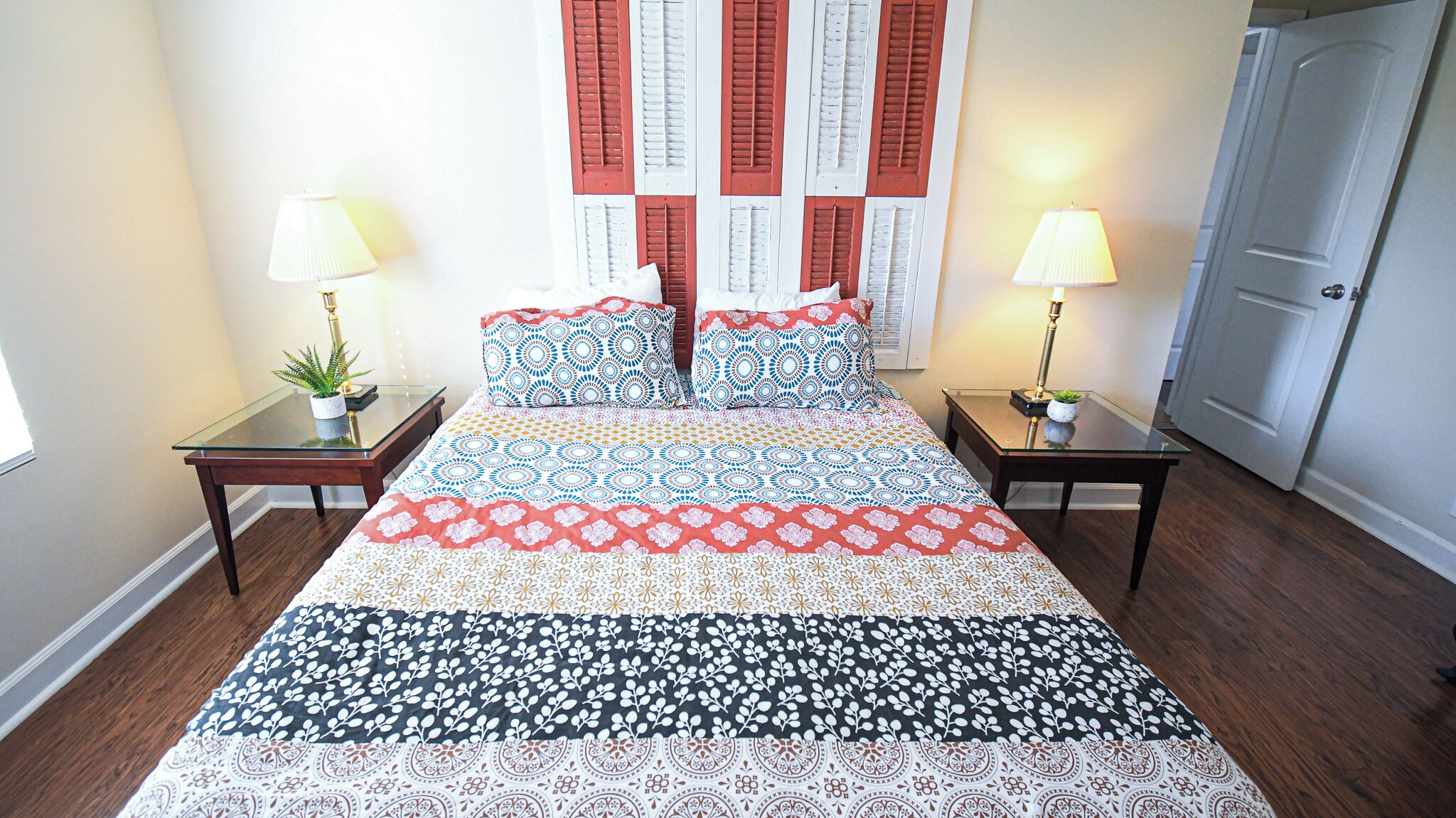 A bed with patterned bedding, two bedside tables with lamps, a potted plant, and a door.