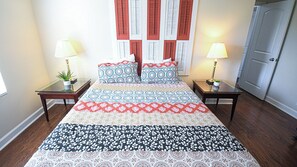 Una cama con ropa de cama estampada, dos mesitas de noche con lámparas, una planta en maceta y una puerta.