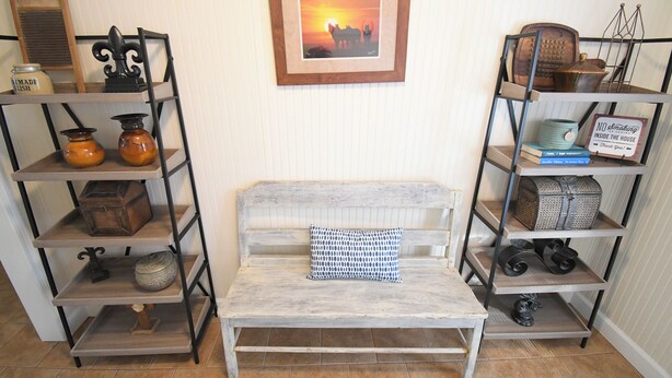 A room with a white-painted bench, a patterned pillow, and shelves with various items including books, jars, and decorative objects.