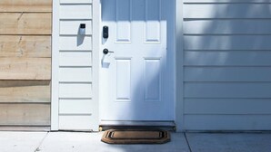 Une porte blanche avec une fente pour le courrier et un trou de serrure, une petite boîte aux lettres et un paillasson sur un chemin de béton menant à un bâtiment aux murs recouverts de bardage en bois et doté d’un toit en métal.