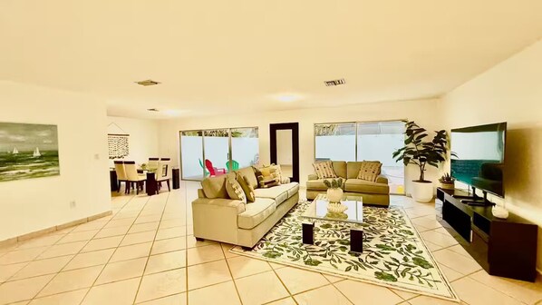 A spacious living room with a sofa, coffee table, and a TV. There is a dining area with chairs and a table in the background.