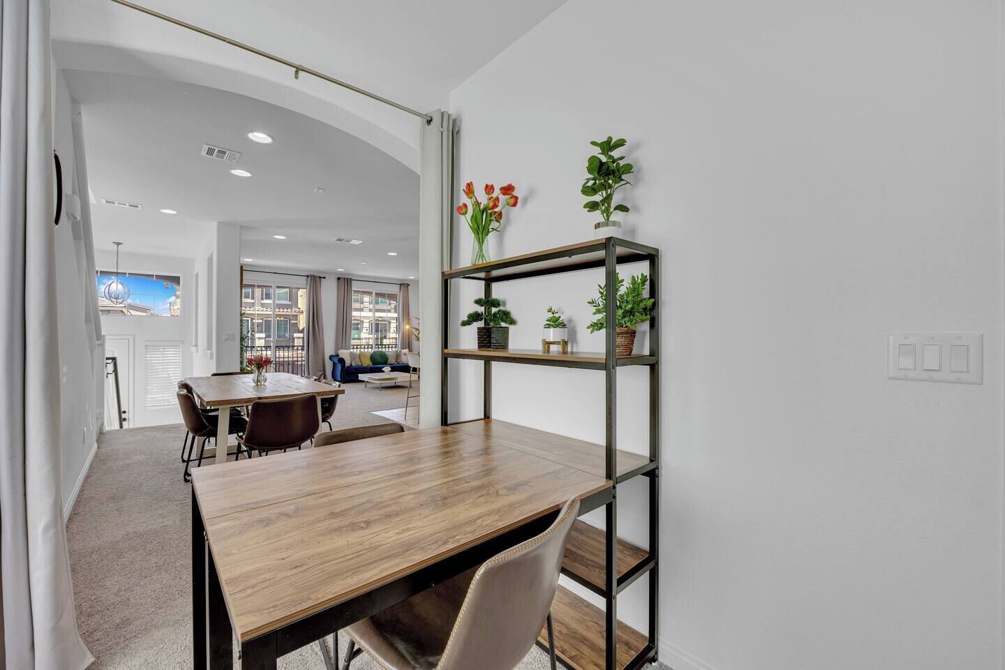A modern interior with a wooden dining table, chairs, a metal shelving unit with plants, and a view of the city through large windows.