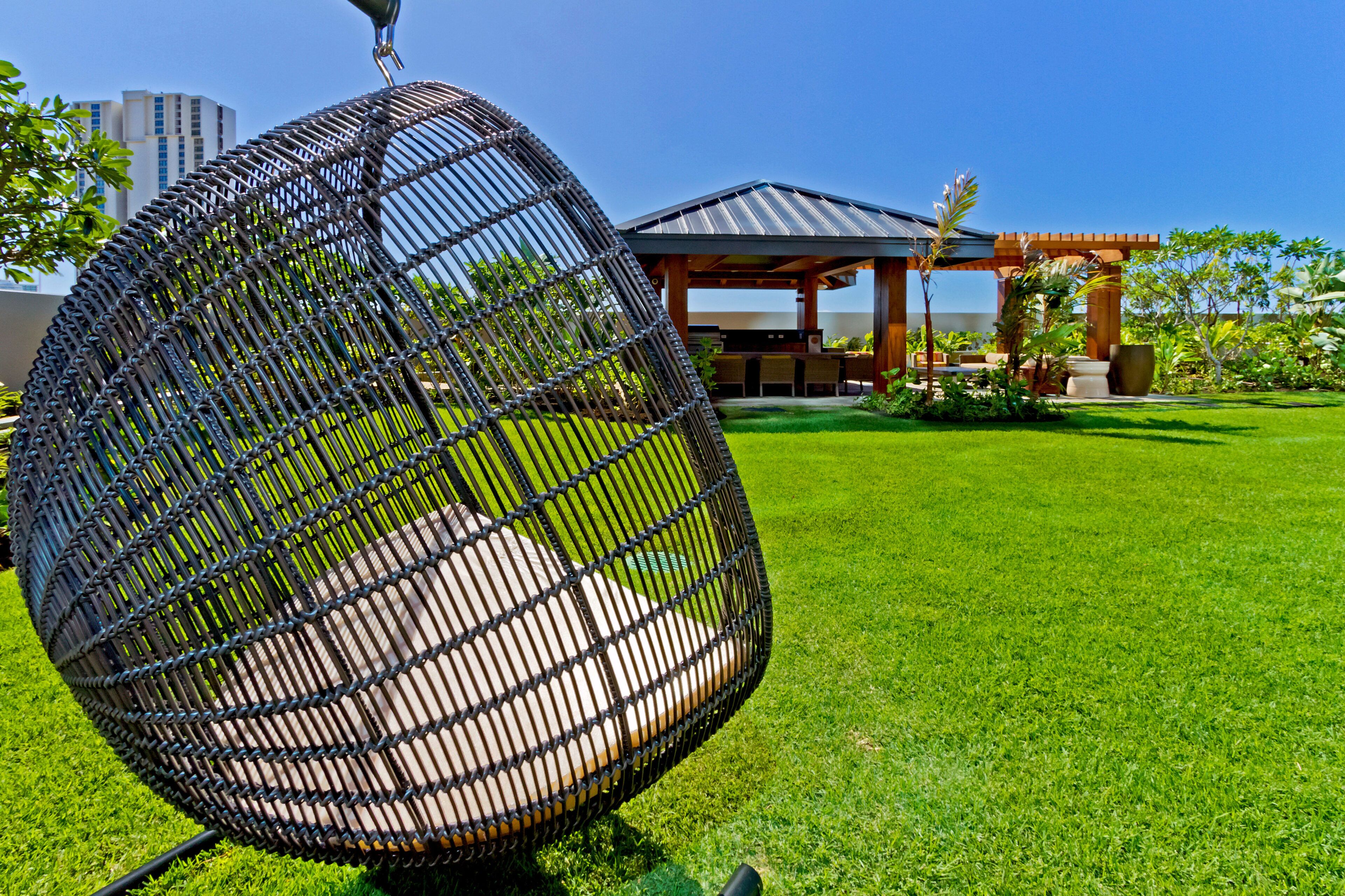 A hanging chair in a well-maintained grassy area with a gazebo and a building in the background.
