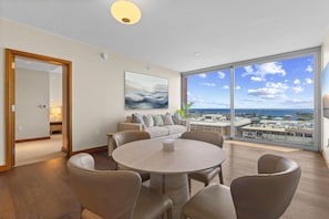 A modern living room with a round table and chairs, a sofa with cushions, a large window with a view of the city and ocean, and a painting on the wall. - Ocean View 2BR/2BA at ONE Ala Moana — With 2 Parking Stalls & Resort Amenities (Honolulu)