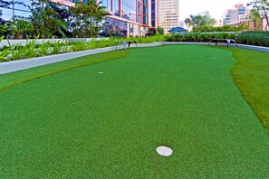 A well-maintained artificial grass golf course with a putting green, surrounded by modern buildings and lush greenery. - Ocean View 2BR/2BA at ONE Ala Moana — With 2 Parking Stalls & Resort Amenities (Honolulu)