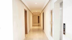 A long, narrow hallway with a polished floor, doors on both sides, and a wall-mounted elevator control panel.