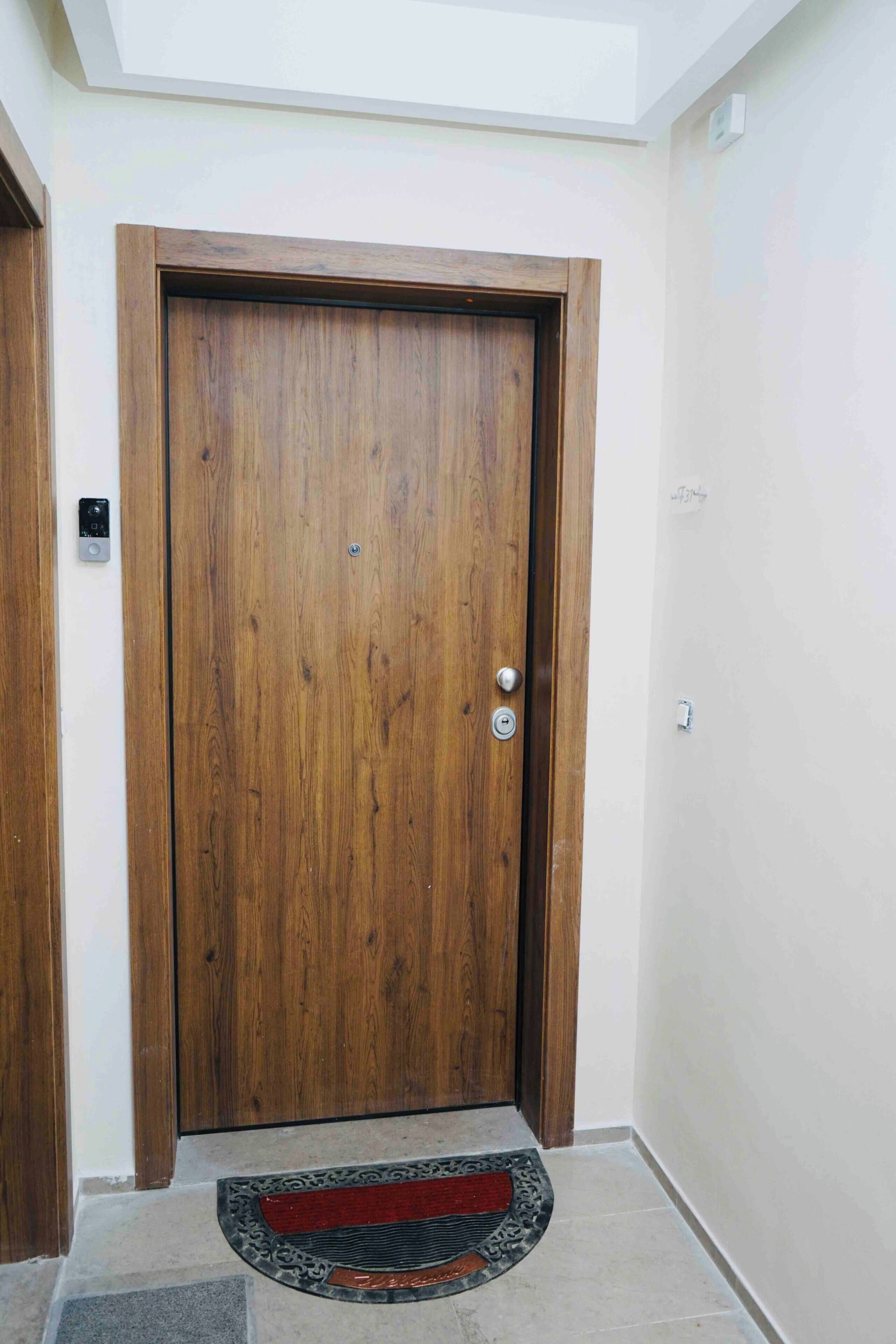 Une porte en bois avec un trou de serrure et une poignée de porte, un petit appareil électronique fixé au mur, et un paillasson aux motifs rouges et noirs à l’entrée.